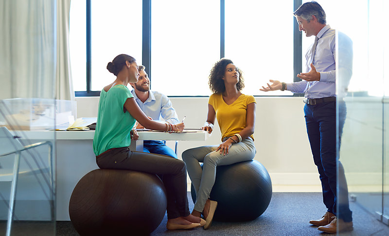 People in a meeting, two of them sitting on an exercise ball rather than a regular office chair. 