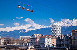 Four cable cars sail over the city of Grenoble against a blue sky and mountains.