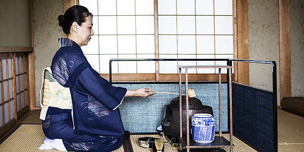 Kneeling Japanese woman in traditional kimono uses a Hishaku (bamboo ladle) during a tea ceremony.