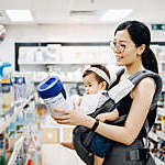 Mother carrying a baby girl in an infant carrier examines tubs of baby formula in a supermarket.