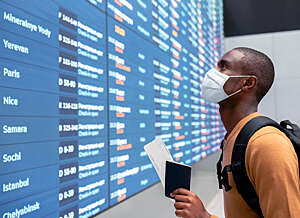 Masked young man, holding a passport and boarding pass, checks his gate number on an airport departures board.