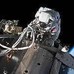 Top view of NASA astronaut Michael Hopkins servicing communications gear on the International Space Station’s Columbus laboratory module, against a backdrop of darkness and blue Earth outline.