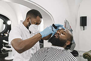 Barber wearing face mask and reusable  gloves doing beard of customer in a barber shop.